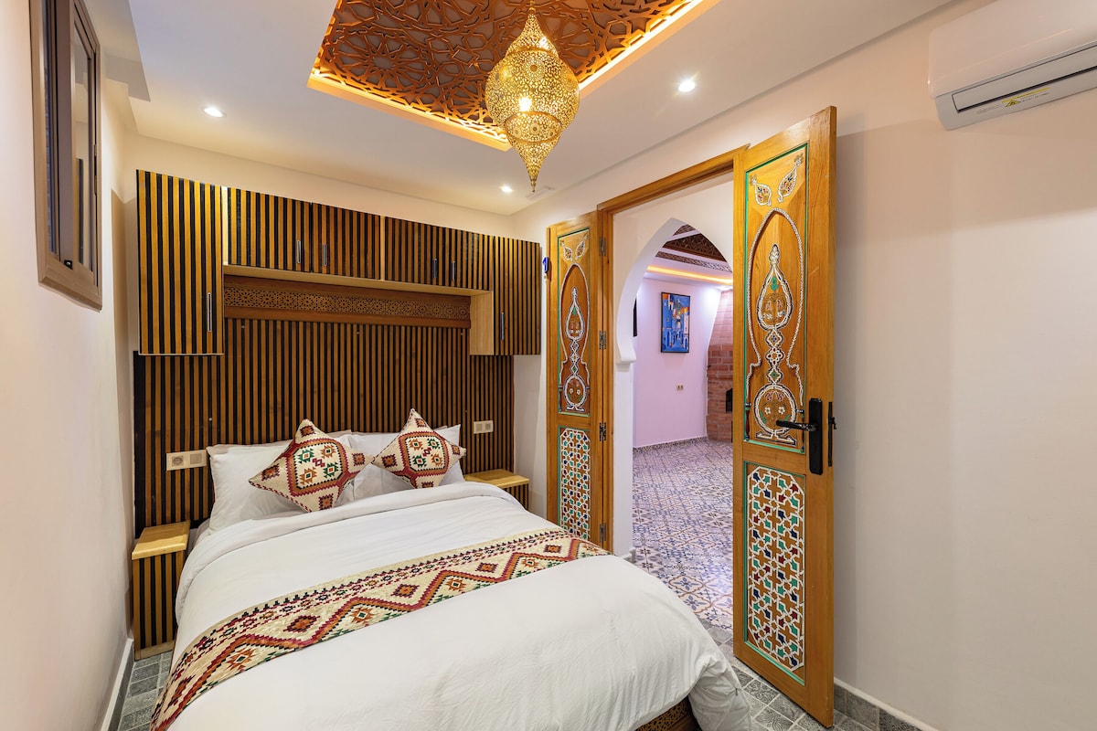 Une chambre à coucher comprenant un lit, une table de chevet, un luminaire décoratif au plafond et une porte ornée de motifs complexes.