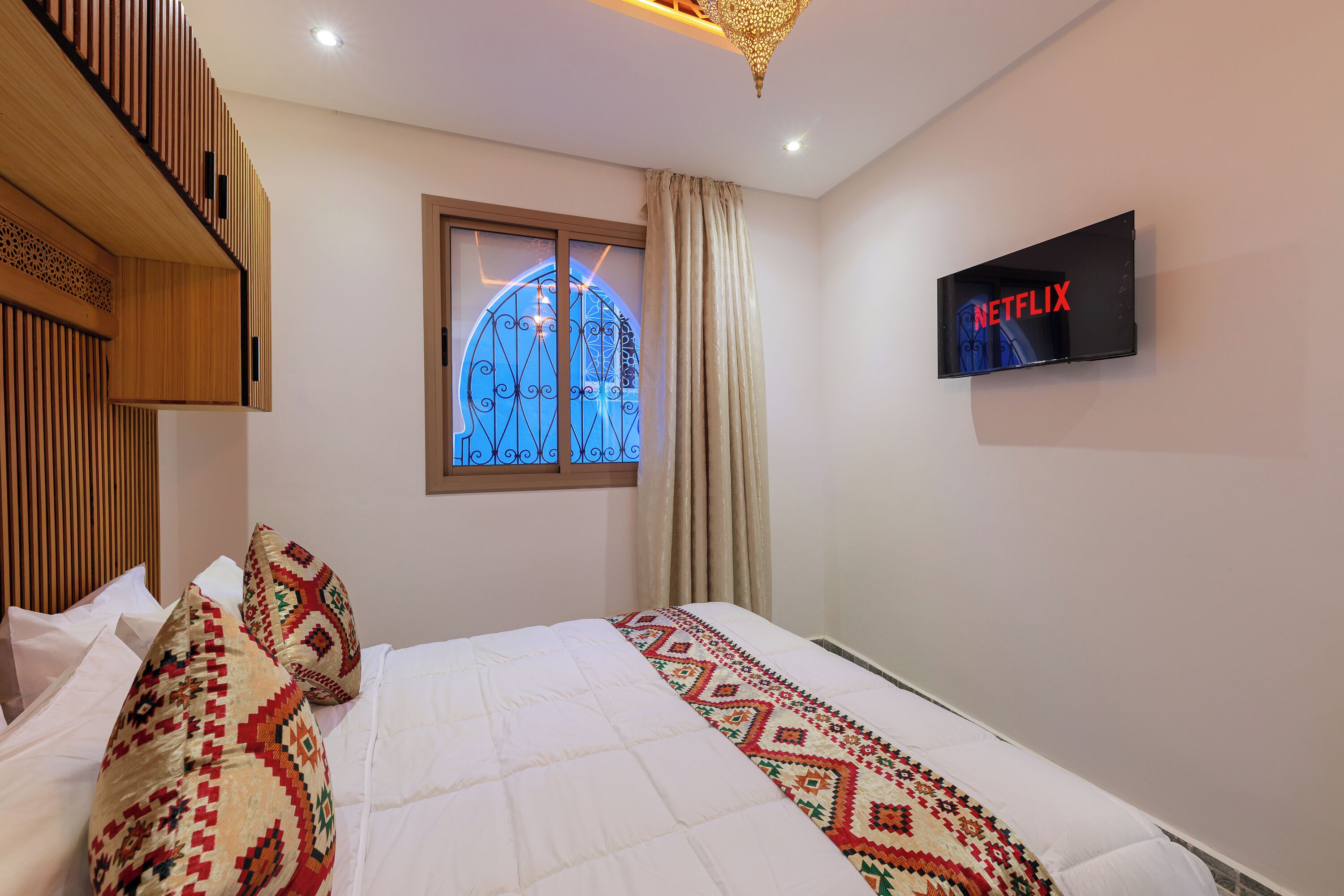 A hotel room with a bed, a TV mounted on the wall, a window with a decorative design, and a ceiling light fixture.
