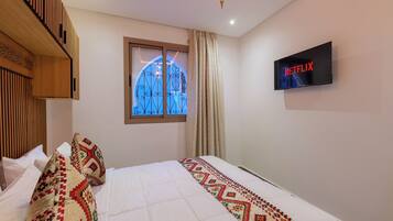 A hotel room with a bed, a TV mounted on the wall, a window with a decorative design, and a ceiling light fixture.