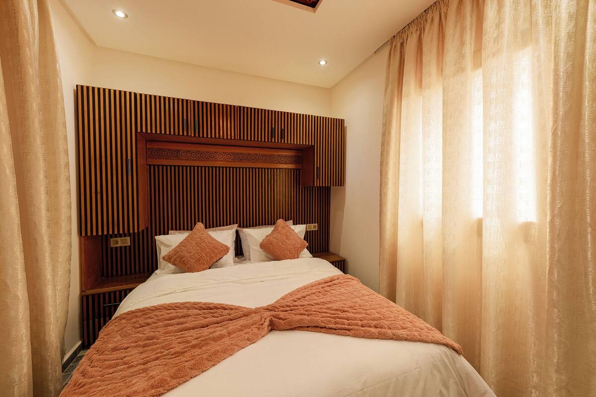A hotel room with a bed, wooden headboard, curtains, and a nightstand.