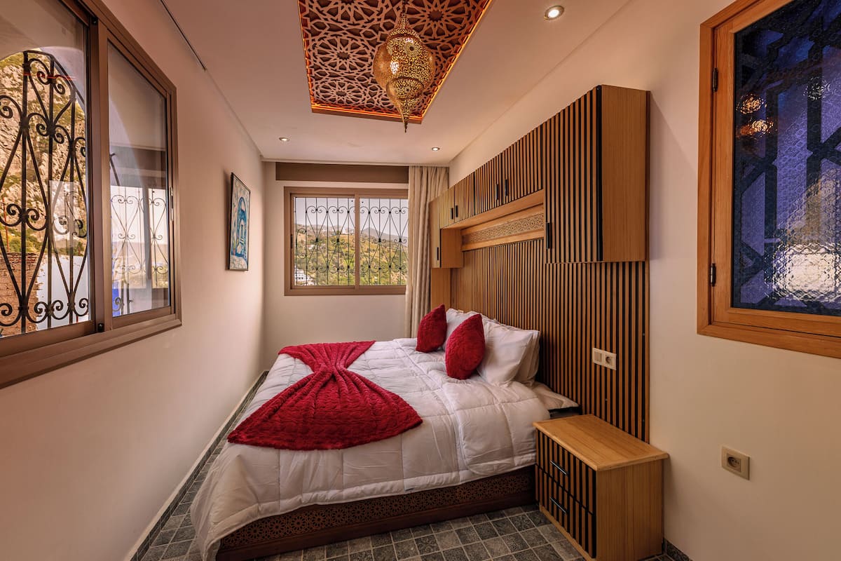 A bedroom with a bed, a window with decorative ironwork, a wooden headboard, and a wall-mounted TV.