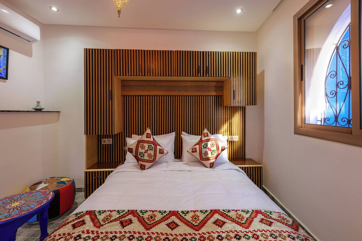 A neatly arranged bedroom with a bed, pillows, a bedside table, a small stool, and a wall-mounted shelf.
