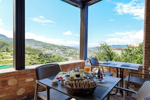 A table set with breakfast items, a bottle of water, and a view of a hilly landscape through large windows. - Riad Seddiki (Chefchaouen)