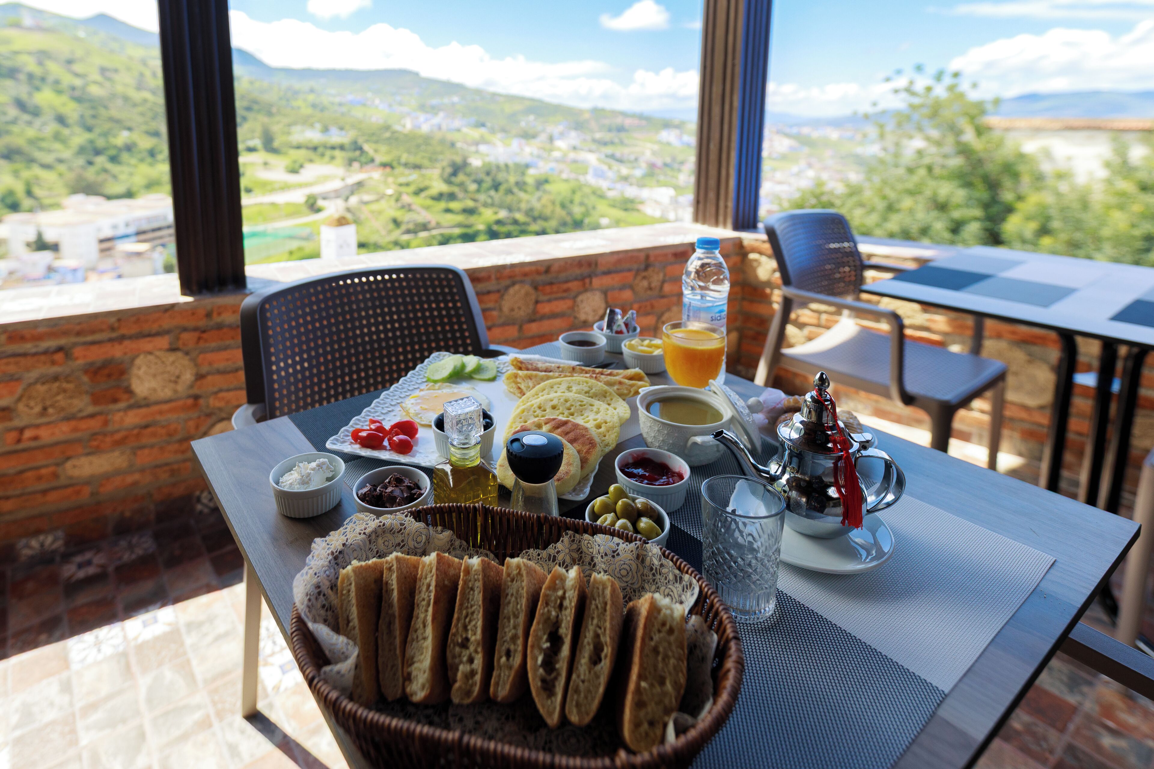 A table set with a variety of food items, including bread, olives, and a beverage, with a scenic view in the background.
