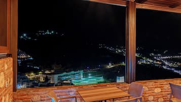 Eine Terrasse mit Holztisch und -stühlen, einer hängenden Lampe und Blick auf eine nächtliche Stadtlandschaft.