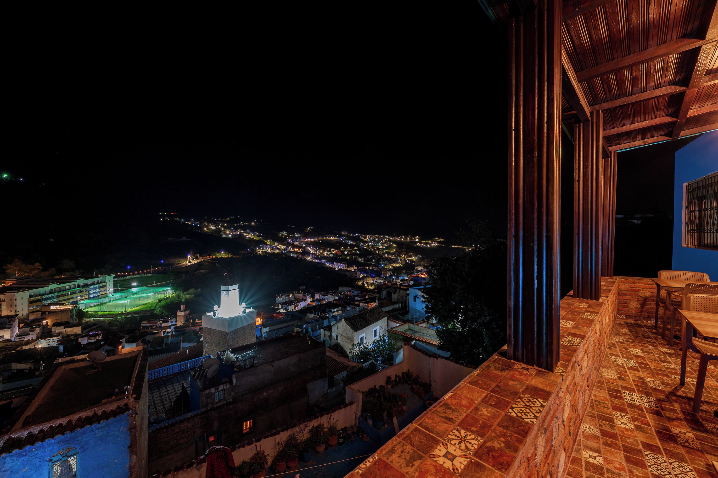 A night view from a balcony with a cityscape in the background, a terrace with a patterned floor, and a view of a lit-up sports field.
