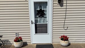 Uma casa com porta branca, uma janela pequena e dois vasos de flores na varanda.