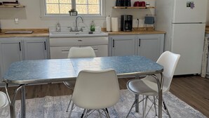 Uma cozinha com uma mesa de jantar, cadeiras, geladeira, uma janela e uma estante com objetos.
