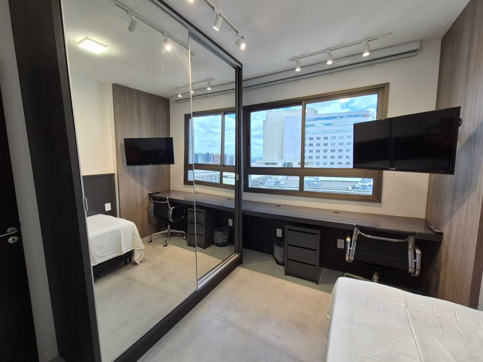 A modern hotel room with a large window, a desk, a chair, a bed, and a flat-screen TV.