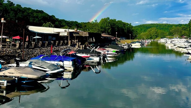 Una marina con barche ormeggiate, un arcobaleno e una collina boscosa sullo sfondo.