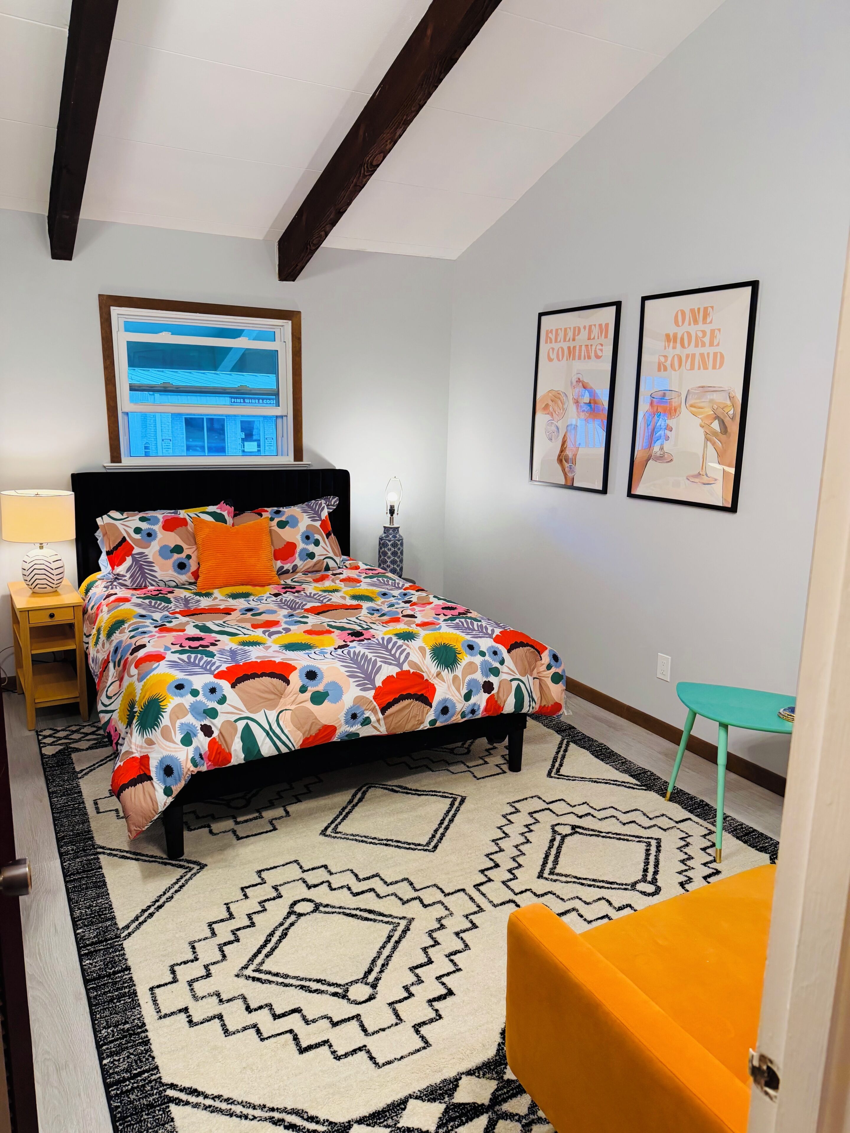 A bedroom with a bed, a nightstand, a lamp, a window, and two framed pictures on the wall.