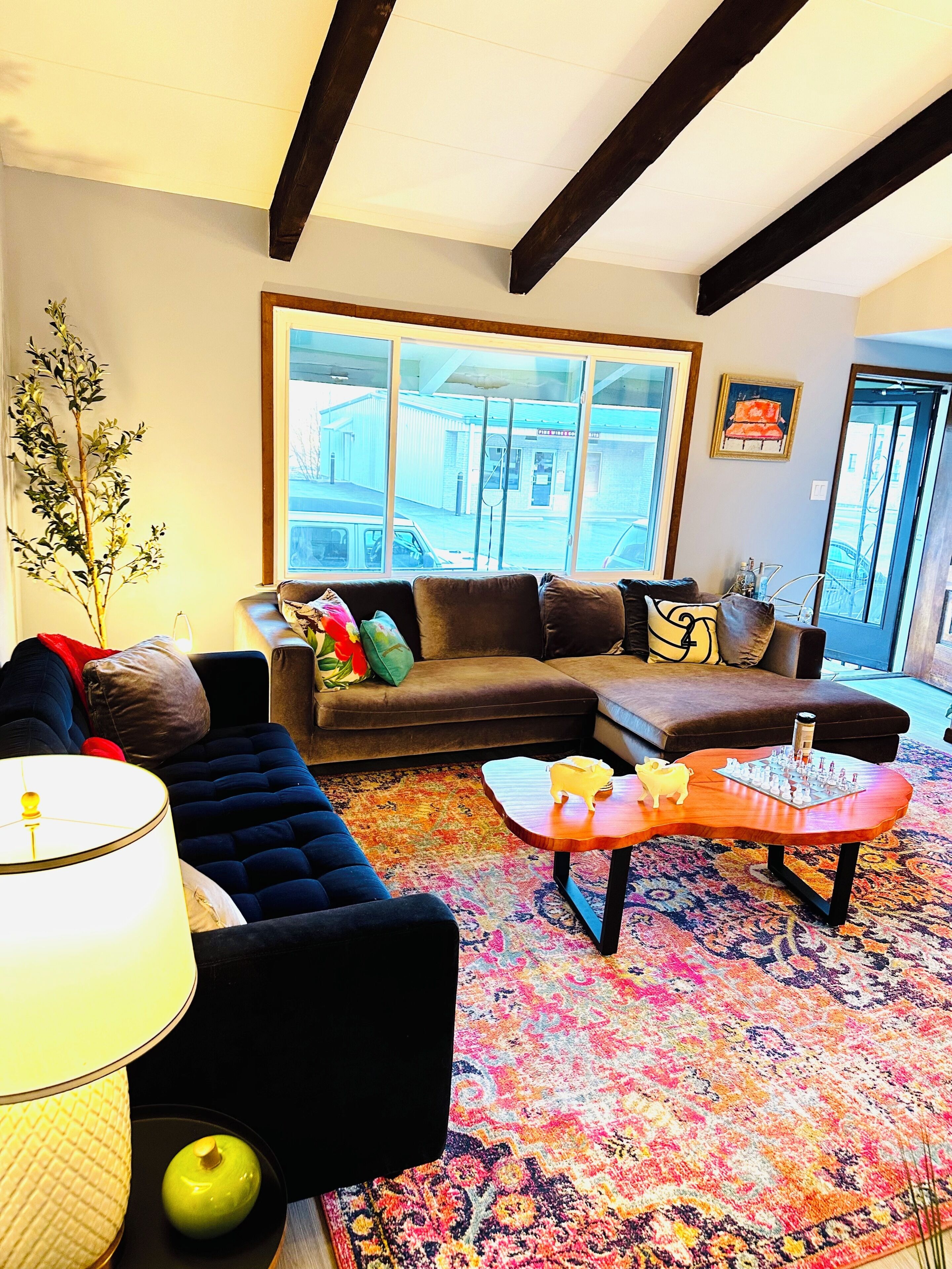 A living room with a sectional sofa, a coffee table, a lamp, and a large window with a view of buildings outside.