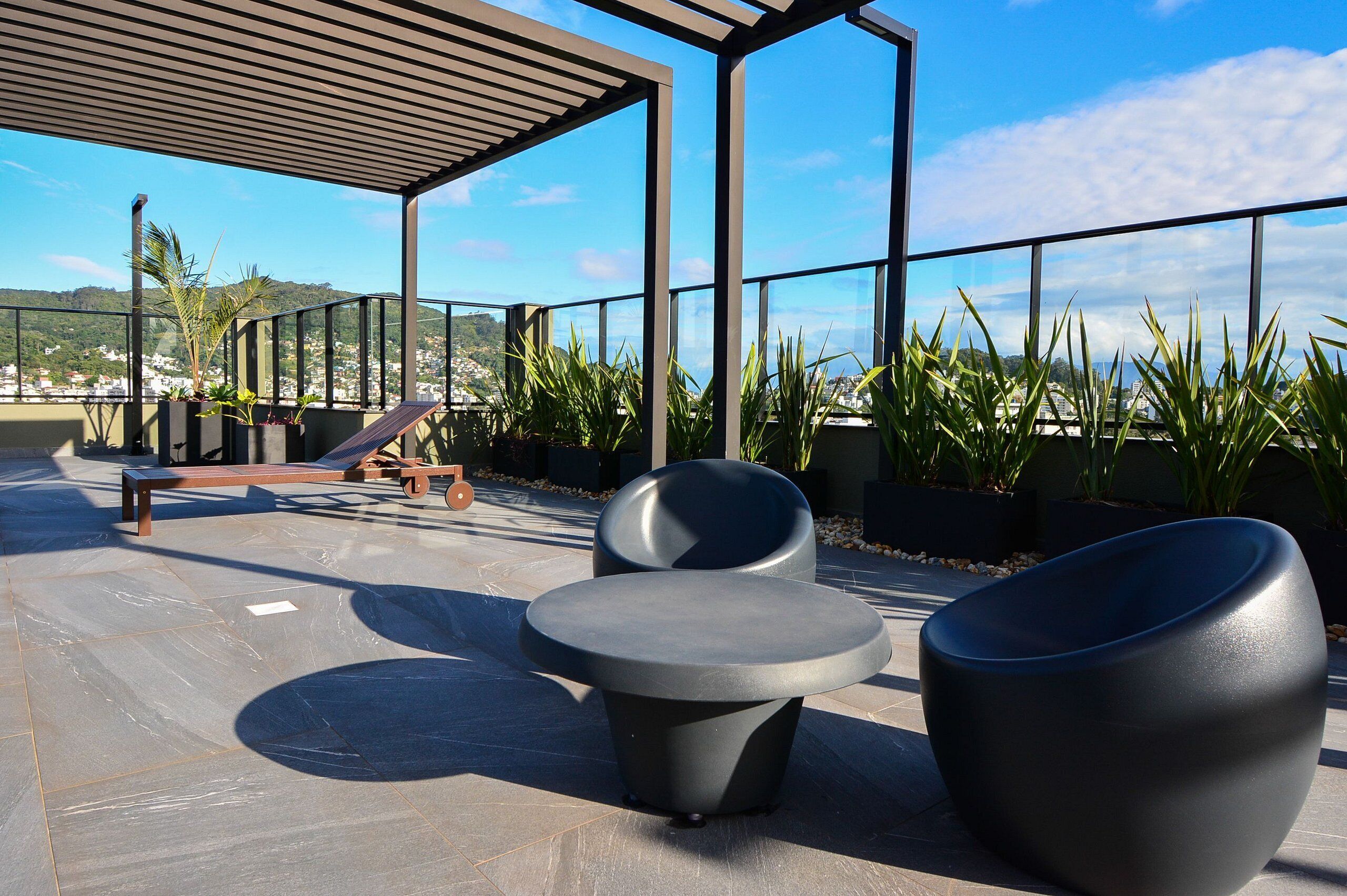 An outdoor seating area with modern chairs and a bench, surrounded by potted plants and a view of a cityscape.