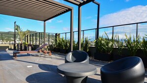 An outdoor seating area with modern chairs and a bench, surrounded by potted plants and a view of a cityscape. - Studio em frente a UFSC e HU - 504R (Florianópolis)