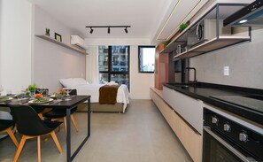 Habitación de hotel pequeña con kitchenette, zona de comedor, cama y un amplio ventanal con vistas a la ciudad.