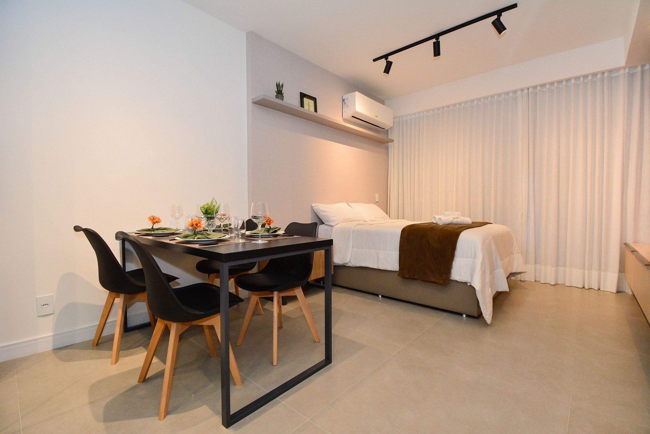 A modern room with a bed, a dining table with chairs, and a small shelf with decorative items.