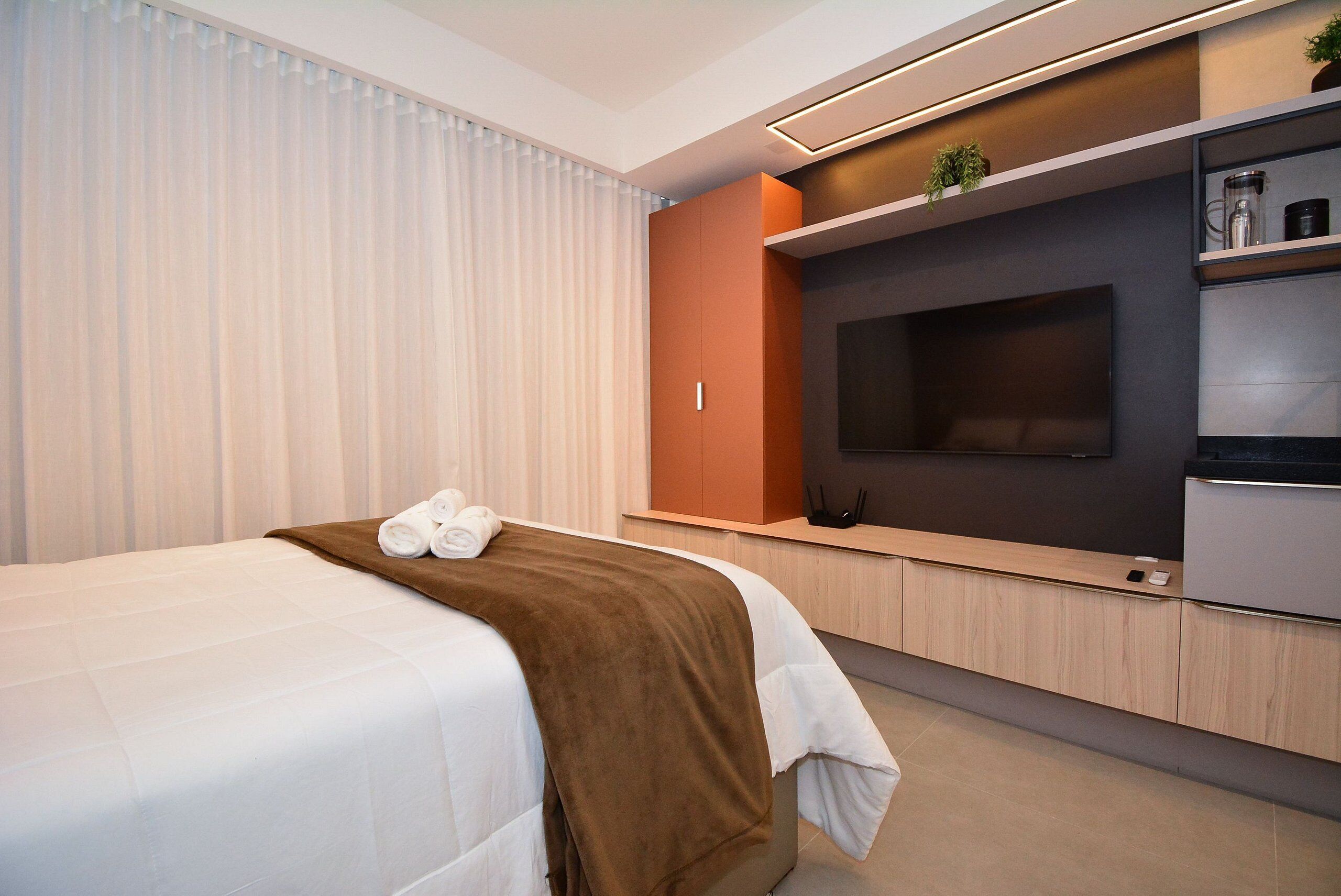 Habitación de hotel moderna con cama, televisor de pantalla plana, cafetera integrada y una pequeña estantería con plantas.