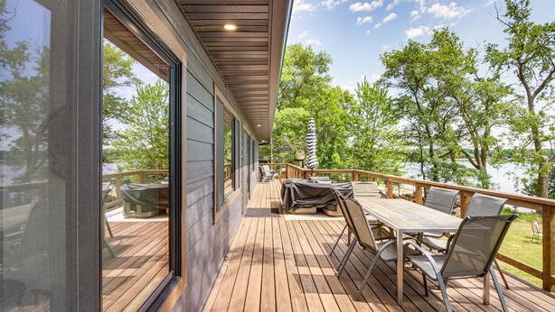 A wooden deck with a table and chairs, overlooking a lake with trees.