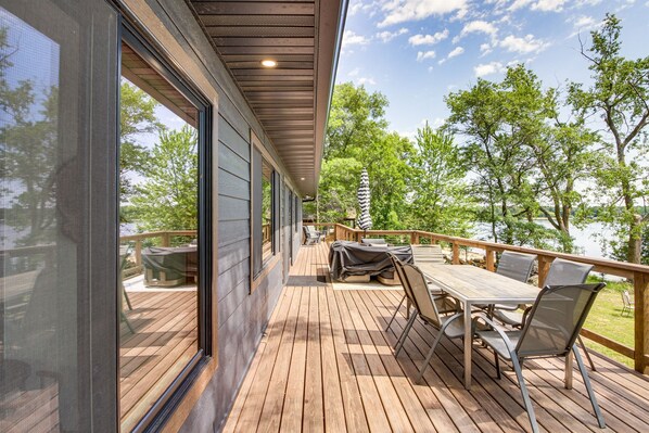A wooden deck with a table and chairs, overlooking a lake with trees.