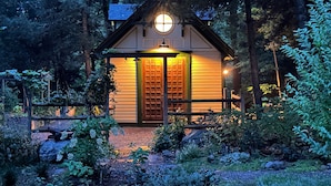 Eine kleine, gemütliche Holzhütte mit einer Holztür und einem Rundfenster, umgeben von dichtem Wald.