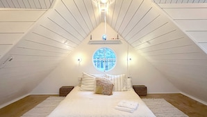 Ein Schlafzimmer mit schrägen Wänden, einem runden Dachfenster, einem Bett mit weißer Bettwäsche und zwei Nachttischen.