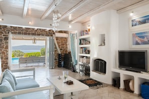 A living room with a stone wall, a fireplace, a TV, and a view of the outdoors. - Holiday Home 'Mykonos' with Sea View, Wi-Fi and Air Conditioning (Άγιοι Θεόδωροι)