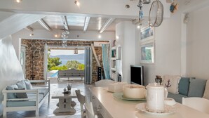 A modern kitchen with a stone wall, a dining area with white furniture, and a view of the outdoors. - Holiday Home 'Mykonos' with Sea View, Wi-Fi and Air Conditioning (Άγιοι Θεόδωροι)
