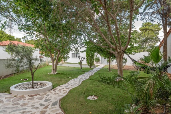 Un jardín con un sendero de piedra, árboles y un edificio blanco con tejado de tejas rojas.