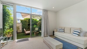 A living room with a sofa, a bench, a TV, and a sliding glass door leading to a garden. - Riverlight Nook - Lake Taupo Holiday Home (Taupo)
