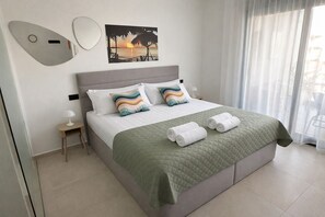A neatly made bed with a green quilt, two pillows, and rolled towels. There is a wall-mounted mirror and a painting of a sunset. - Casa Anna - Velapi II - Los Alcázares (San Javier)