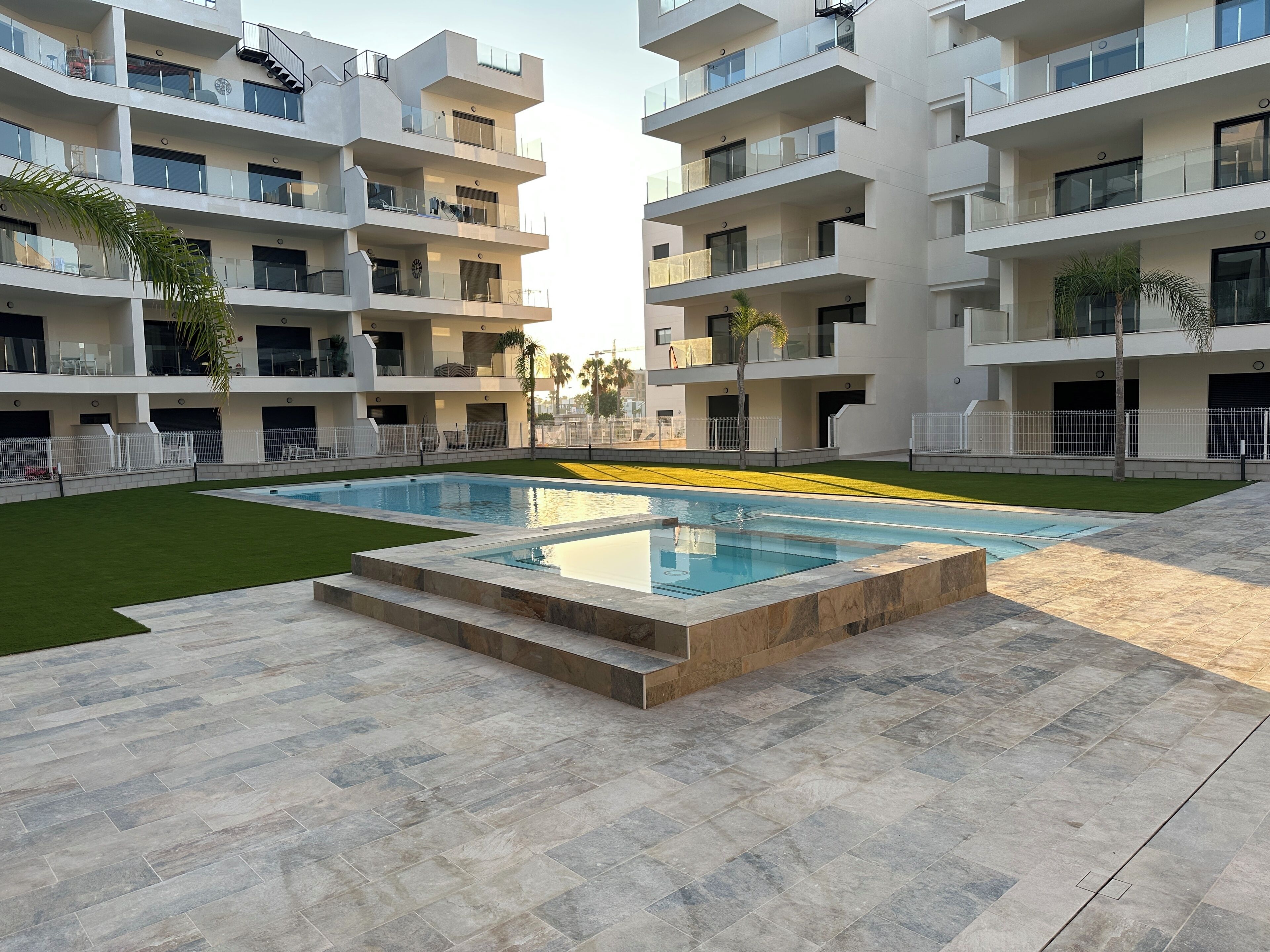Ein modernes, mehrstöckiges Wohngebäude mit einem zentralen Schwimmbecken, umgeben von gepflegten Rasenflächen und einem gepflasterten Gehweg.