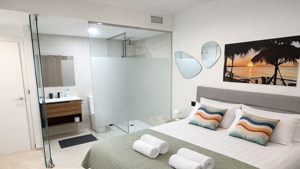 A modern hotel room with a glass shower enclosure, a bed with two pillows, and a sunset painting on the wall. - Casa Anna - Velapi II - Los Alcázares (San Javier)