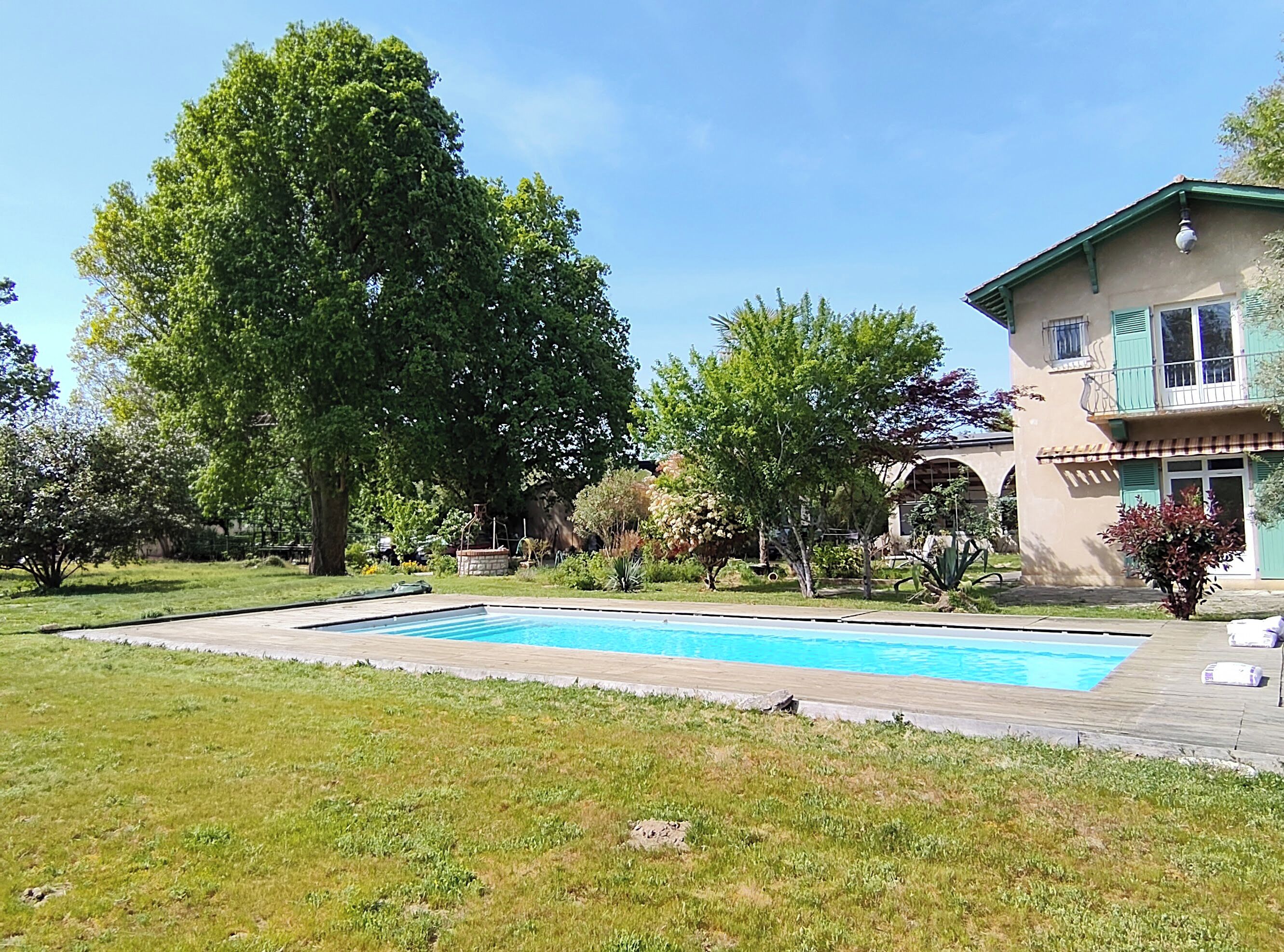 A swimming pool surrounded by a grassy area, a large tree, and a building with a balcony.