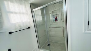Casa de banho com cabine de duche em vidro, porta-toalhas, suporte de papel higiénico e um pequeno estante com artigos de higiene pessoal.