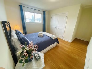 A bedroom with a bed, a nightstand, a lamp, a window with curtains, a door, and a view of houses outside.