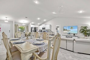 Una cocina moderna con armarios blancos, un comedor con una mesa dispuesta para seis personas y una sala de estar con sofá y televisor.