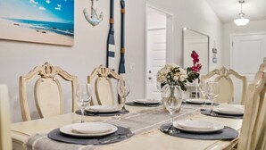 A dining area with a table set for four, chairs with ornate backs, a vase with flowers, and a painting of a beach scene on the wall.