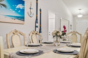 A dining area with a table set for four, chairs with ornate backs, a vase with flowers, and a painting of a beach scene on the wall. - NEW Luxury Build + Private Saltwater Pool & Spa (Venice)