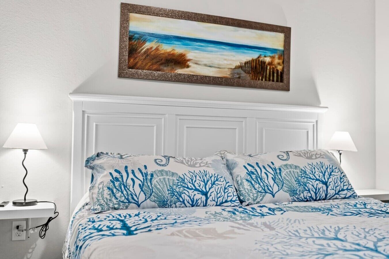 Ein Schlafzimmer mit weißem Kopfteil, einem Bild von einer Strandlandschaft und einem Bett mit blau-weißer Bettwäsche.