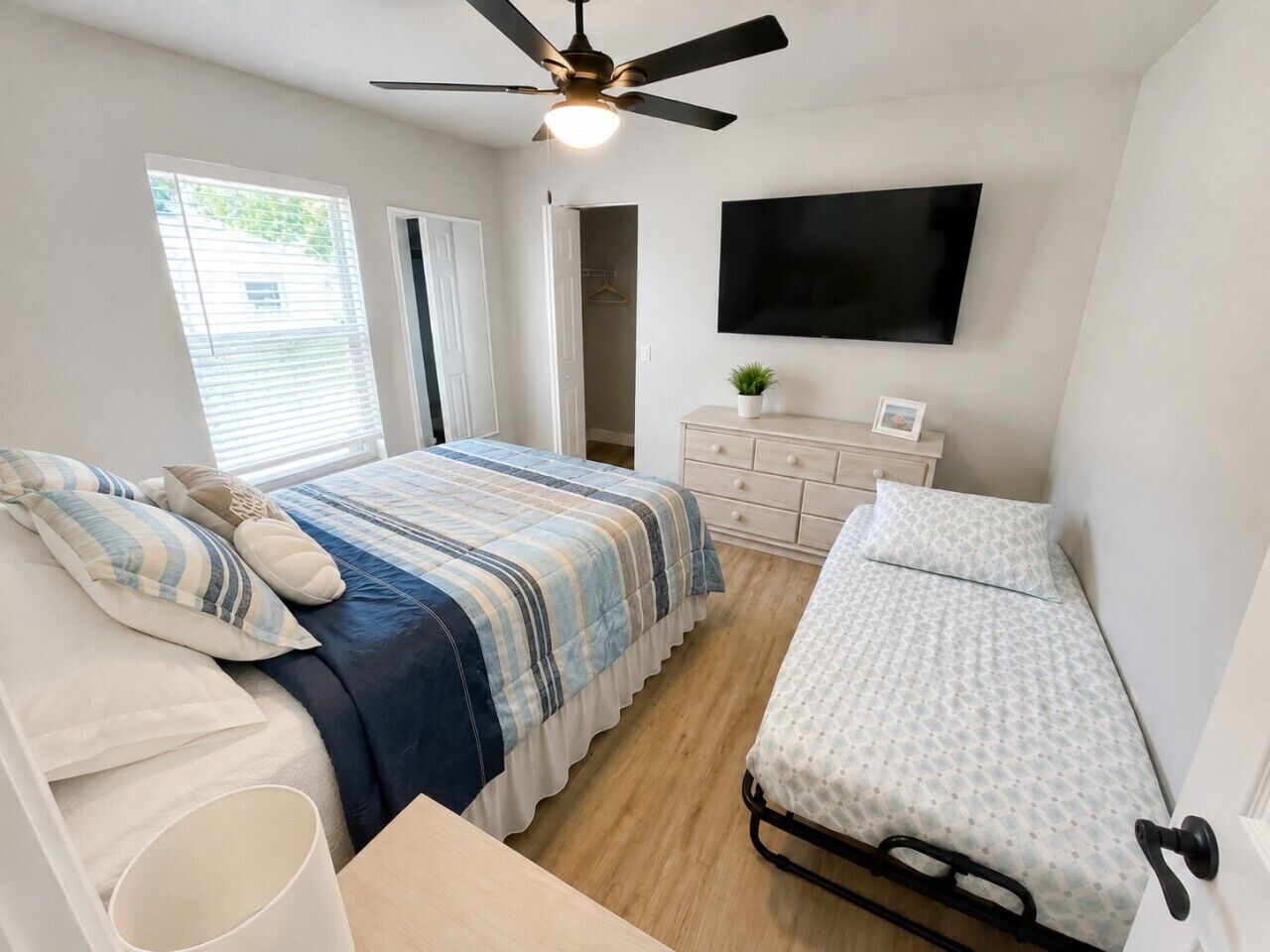 A bedroom with a bed, a bedside table, a dresser, a ceiling fan, a TV, and a window.