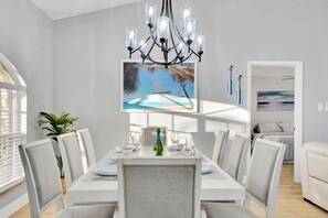 Zona pranzo con tavolo bianco, sedie grigie, un lampadario e un quadro raffigurante una spiaggia.