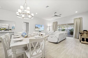 Un comedor moderno y bien iluminado con mobiliario blanco, una araña y vistas al exterior a través de amplias ventanas.