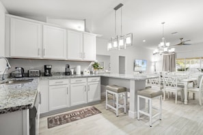 Une cuisine moderne avec des meubles blancs, des plans de travail en granit, un îlot central doté de tabourets et un coin repas avec des chaises blanches.