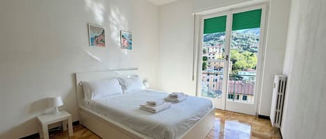 Une chambre à coucher avec un lit, une table de chevet, une lampe, un radiateur et une vue sur des immeubles par la fenêtre.