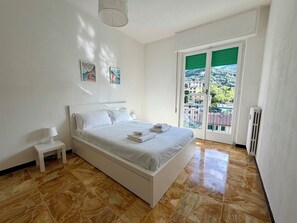 Ein Schlafzimmer mit Bett, Nachttisch, Lampe, Heizkörper und Blick auf Gebäude durch ein Fenster.