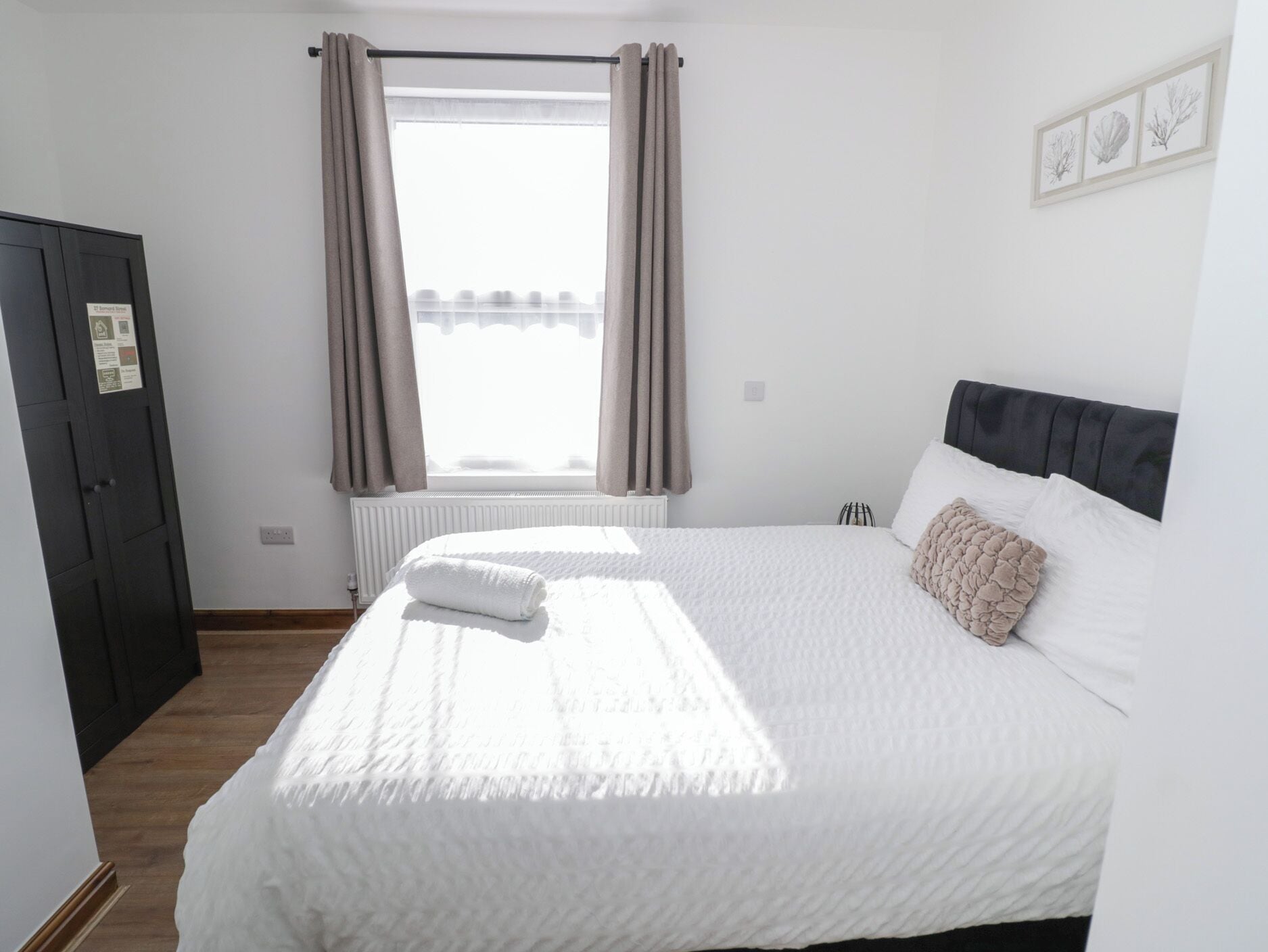A bedroom with a bed, a radiator, a window with curtains, a door, and a framed picture on the wall.