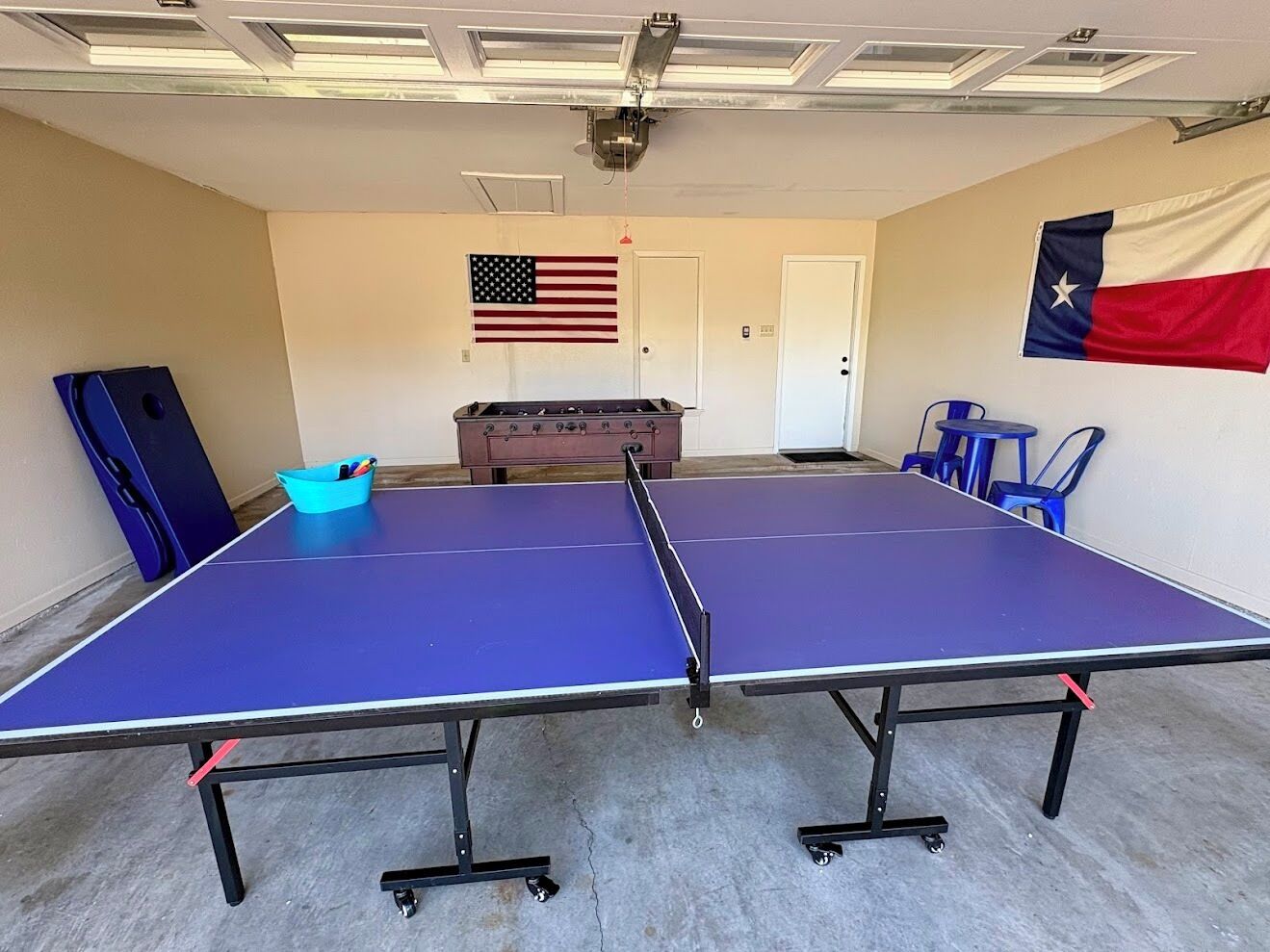 Una mesa de ping pong con dos sillas, un cubo azul y dos banderas en la pared.