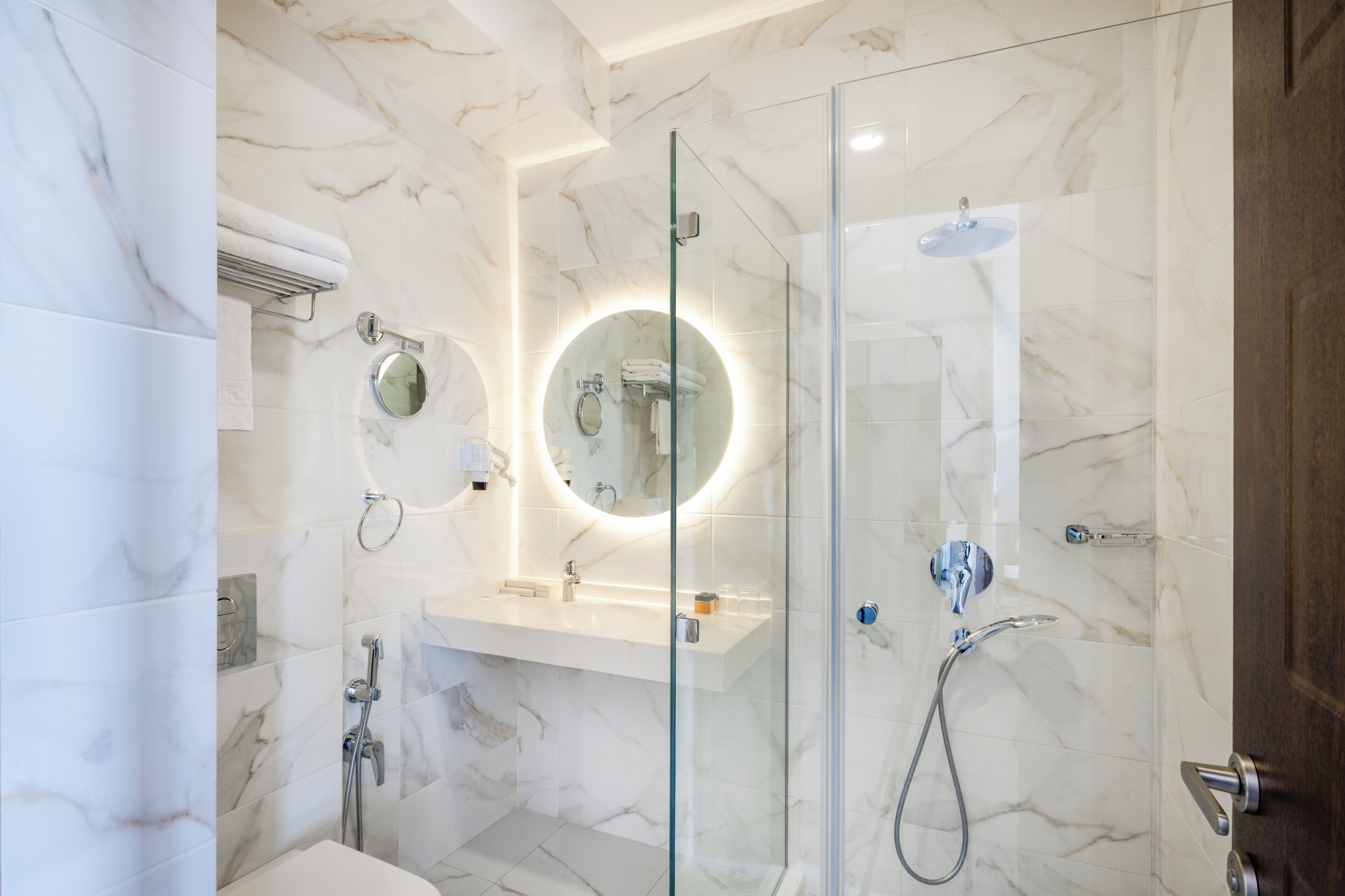 Une salle de bain moderne avec une douche à parois de verre, un miroir rond et un mur recouvert de carreaux de marbre.