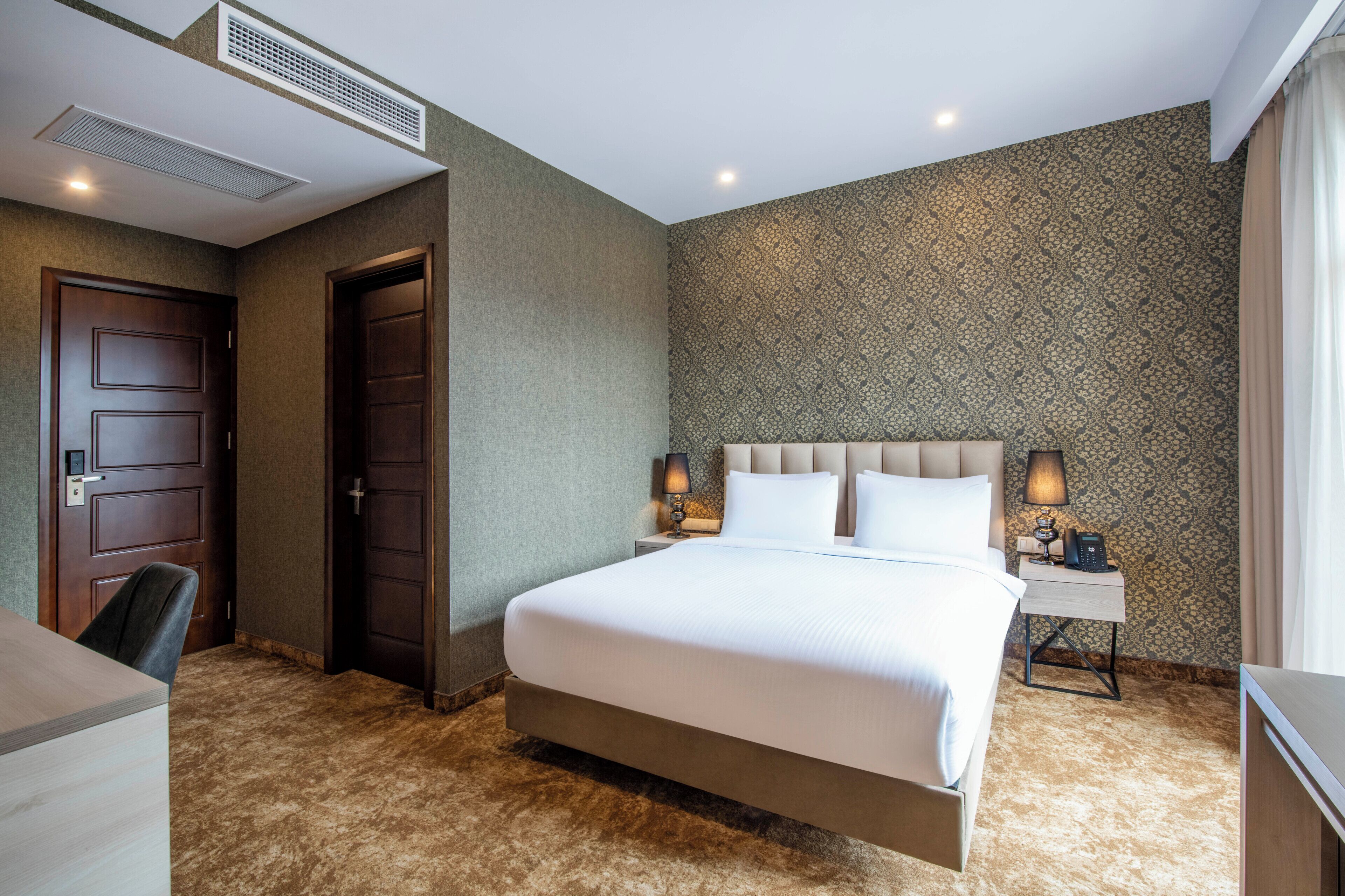 Habitación de hotel con una cama grande, un escritorio, una silla, un armario, una puerta, una mesita de noche con una lámpara y un papel tapiz estampado.