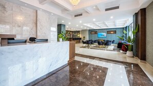 Un hall d’hôtel avec un comptoir de réception en marbre, un lustre, un espace repas avec des tables et des chaises, et des plantes en pot.